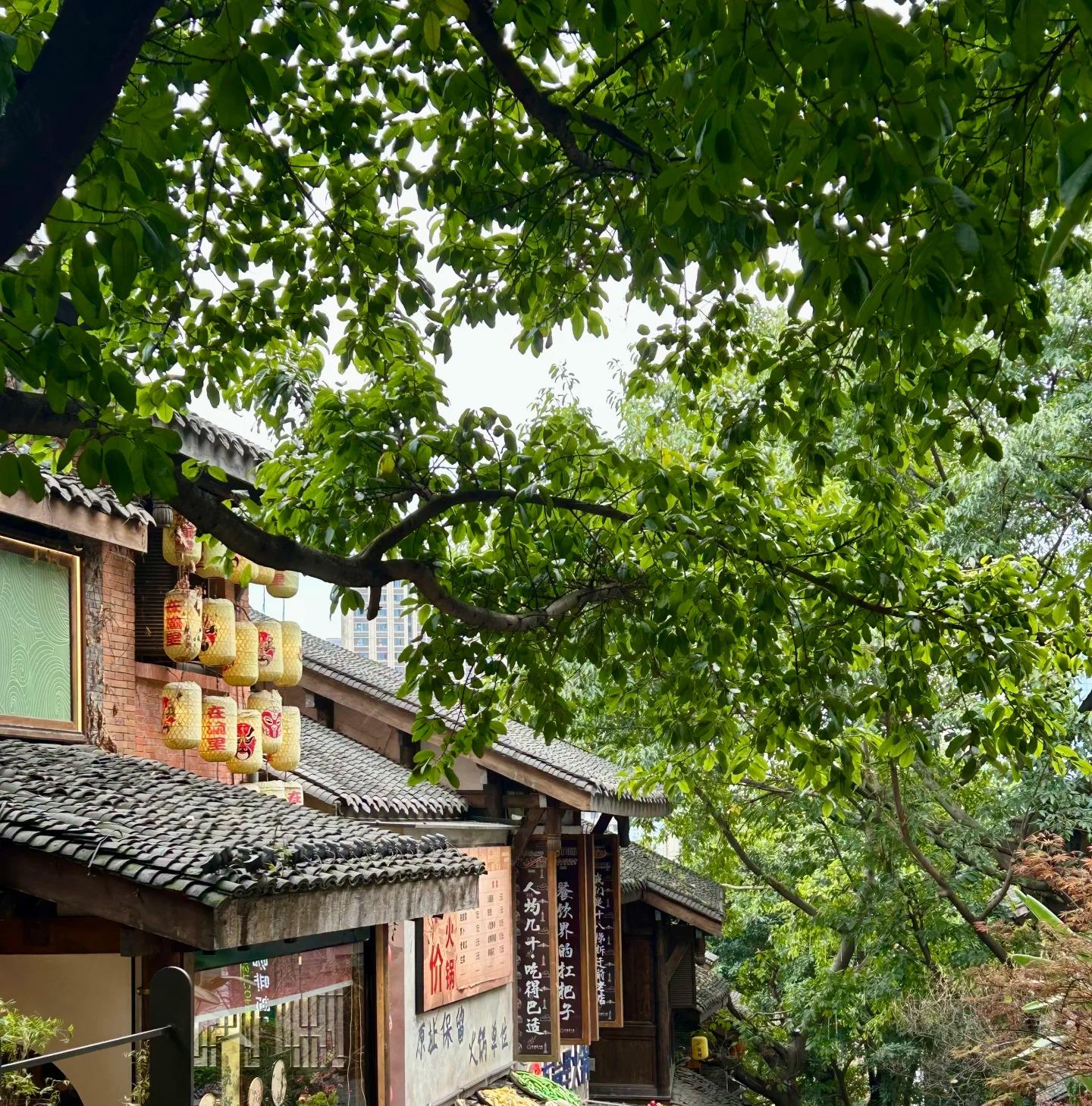 Painted opera-mask lanterns hanging from a brick building on an old street, with hotpot restaurant banners below and trays of ingredients displayed on a tiled roof.