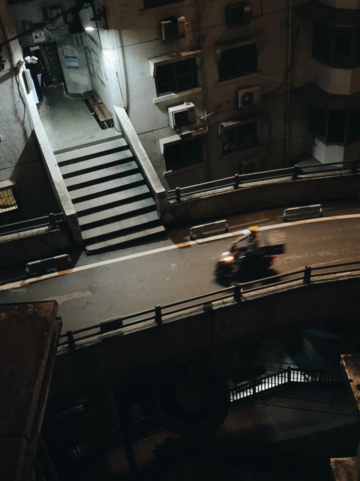 Night view looking down onto a curving road with a motorcyclist, a flight of steps descending from it to a lower level, and apartment windows behind.