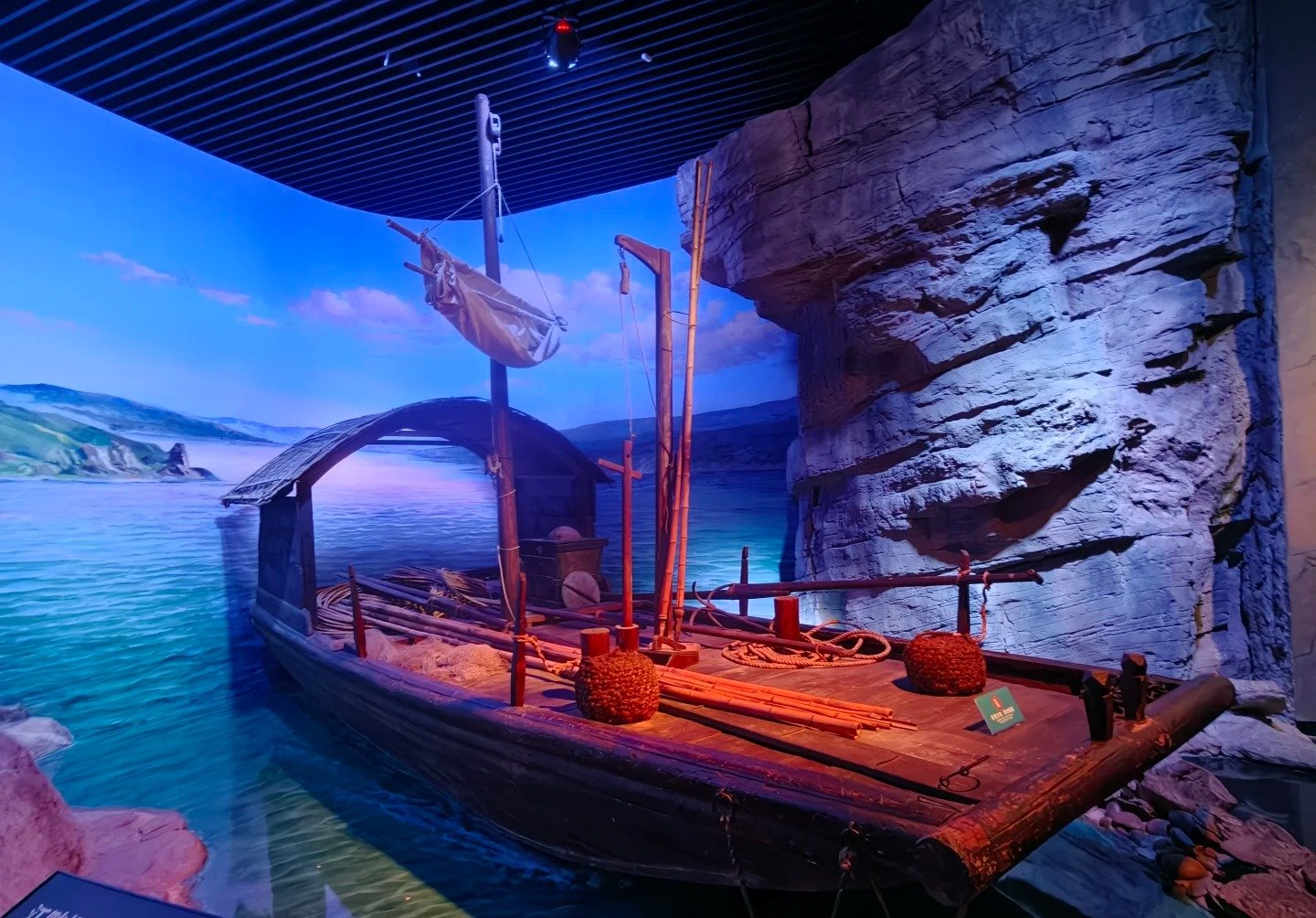 A full-size reconstructed wooden Yangtze cargo boat with mast and coiled ropes, set against a painted gorge backdrop and simulated water inside the Majestic Three Gorges gallery.