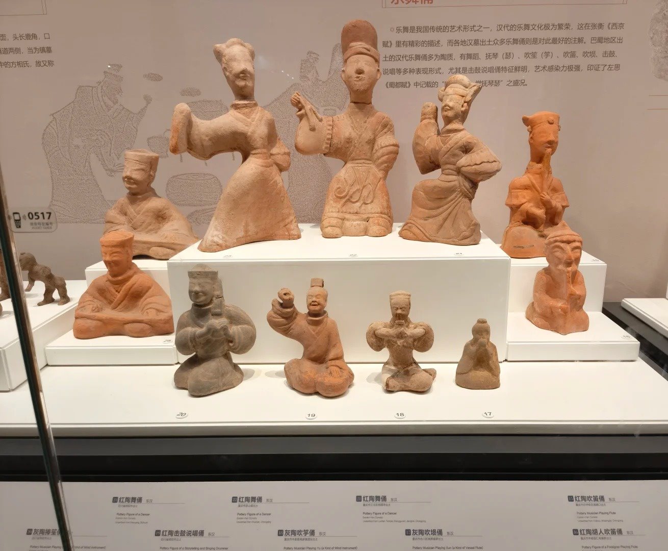 A museum case of reddish Han-dynasty pottery figures of dancers and musicians, above a row of wall labels printed in Chinese and English giving each figure's name, dynasty and find site.