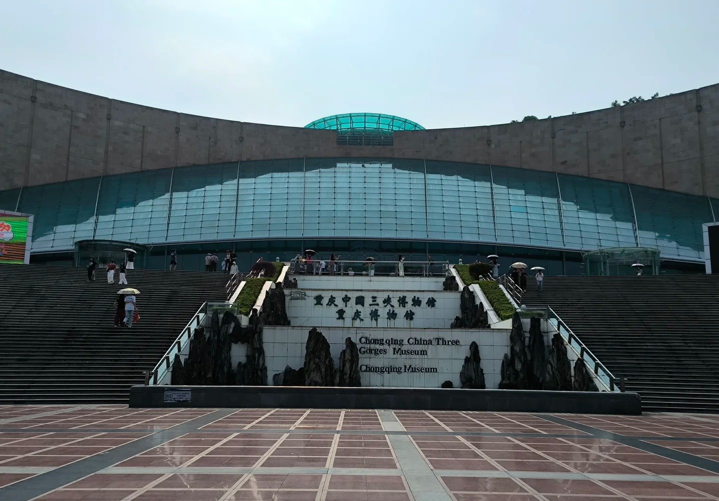 The museum's stone name wall at the foot of its steps, carved with 重庆中国三峡博物馆 and 重庆博物馆 above the English lines Chongqing China Three Gorges Museum and Chongqing Museum, below the curved stone facade and teal glass frontage.