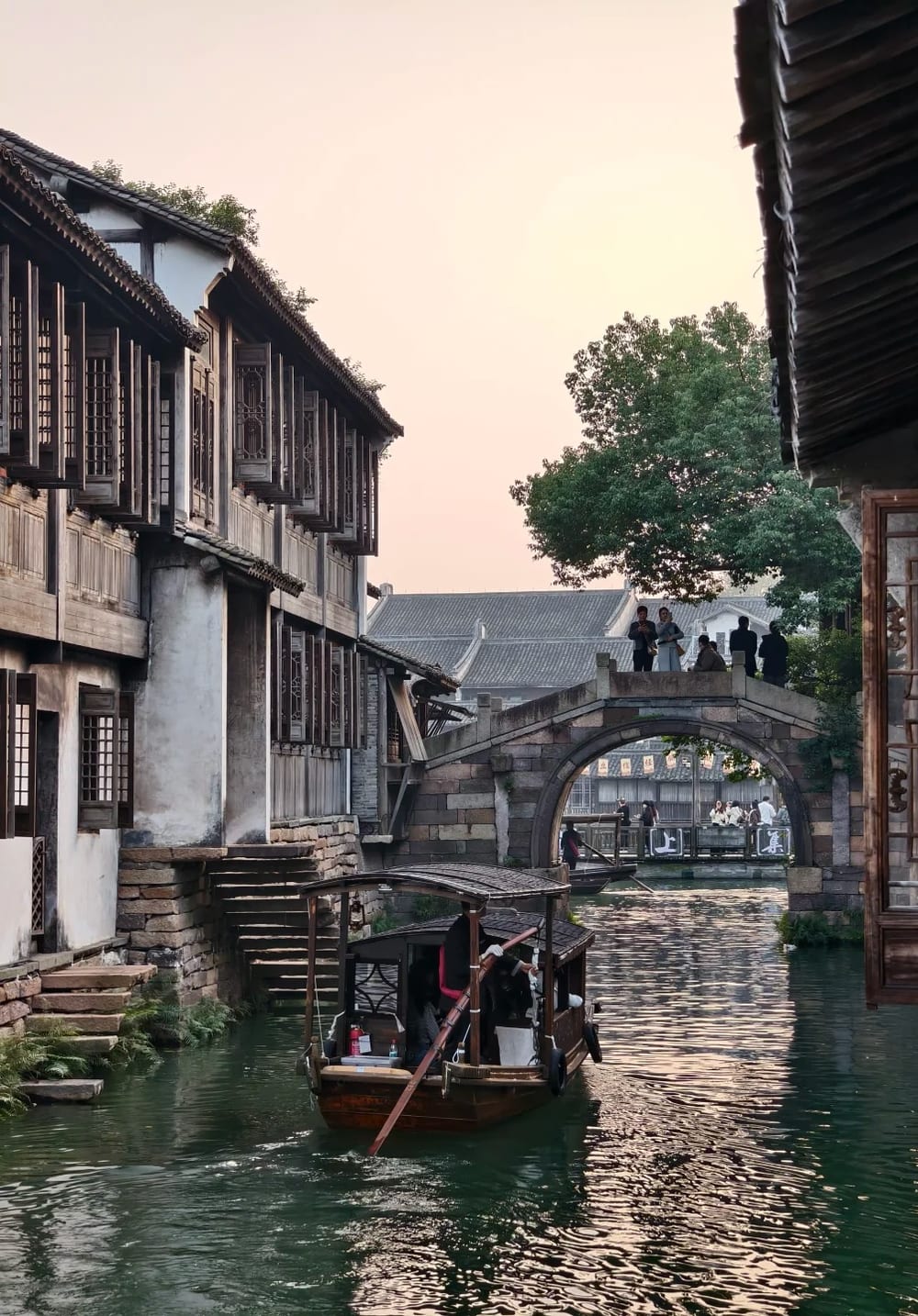 A hand-poled wooden boat on a Wuzhen canal at dusk, the boatman standing at the stern working a long yuloh oar, with a stone arch bridge behind and lit signs visible through its arch.
