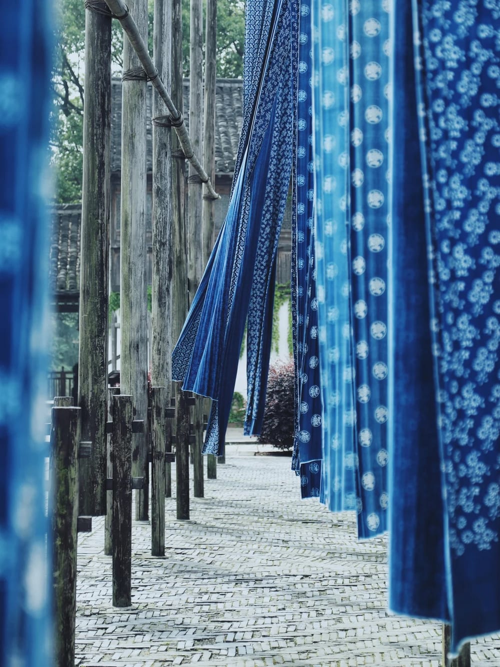 Rows of tall weathered timber poles in a Wuzhen dye works, hung with long lengths of blue-and-white indigo-printed cloth drying above a brick-paved lane.