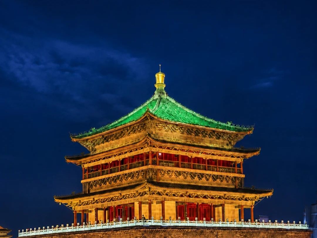The Bell Tower of Xi'an floodlit at night against a deep blue sky, its green-tiled pyramid roof and gold finial lit from below.