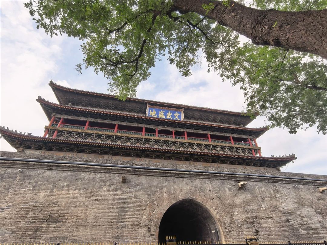 The Drum Tower seen from below, its blue and gold plaque reading wen wu sheng di, above a grey brick base with a large open archway.