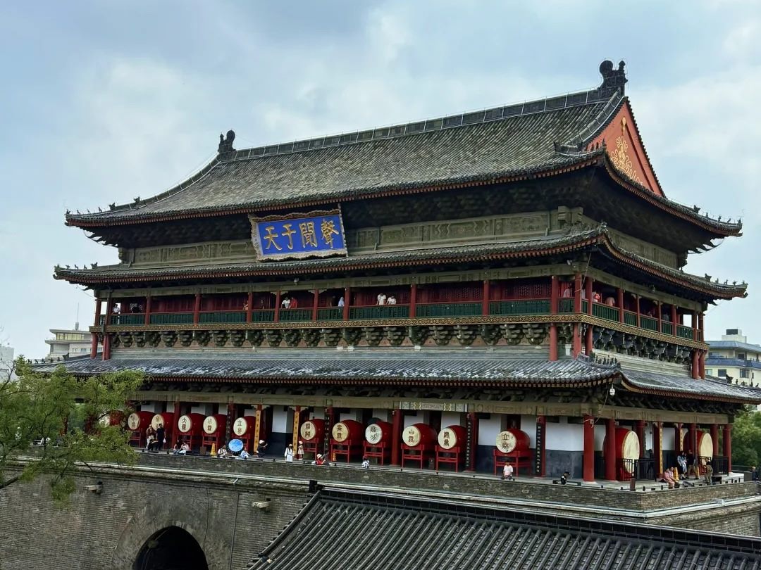 The Drum Tower's lower gallery lined with large red drums, each with a gold-lettered head, beneath a blue plaque reading sheng wen yu tian.