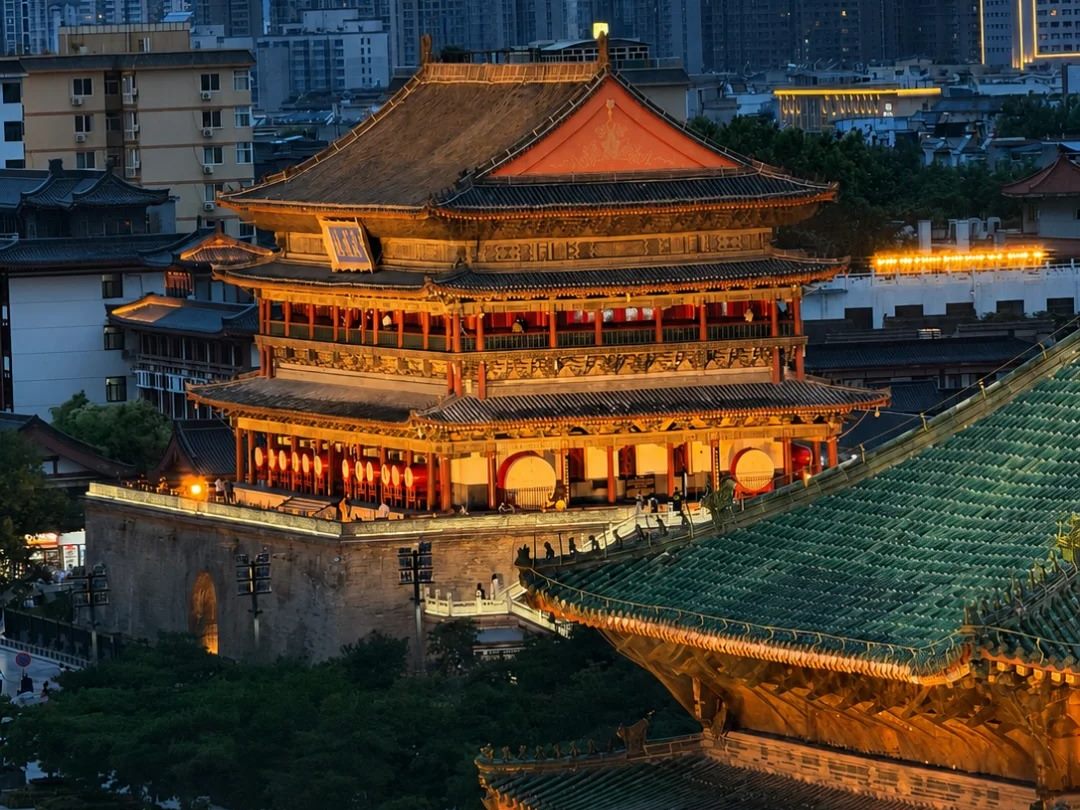 Blue-hour view over central Xi'an with the floodlit Drum Tower in the middle distance and the green-tiled pyramid roof of the Bell Tower in the lower right foreground.