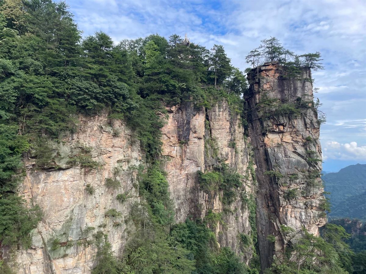A sheer quartz-sandstone wall face in Yangjiajie topped by a small clifftop pavilion, with a free-standing pillar beside it, Zhangjiajie National Forest Park.