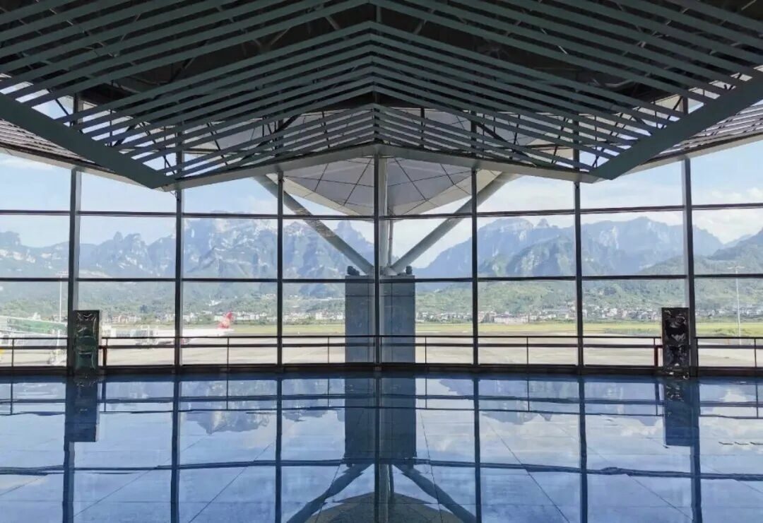 The view through the glass curtain wall of Zhangjiajie Hehua Airport's terminal: an aircraft on the apron, and a wall of jagged sandstone-quartzite ridges rising directly behind the runway.